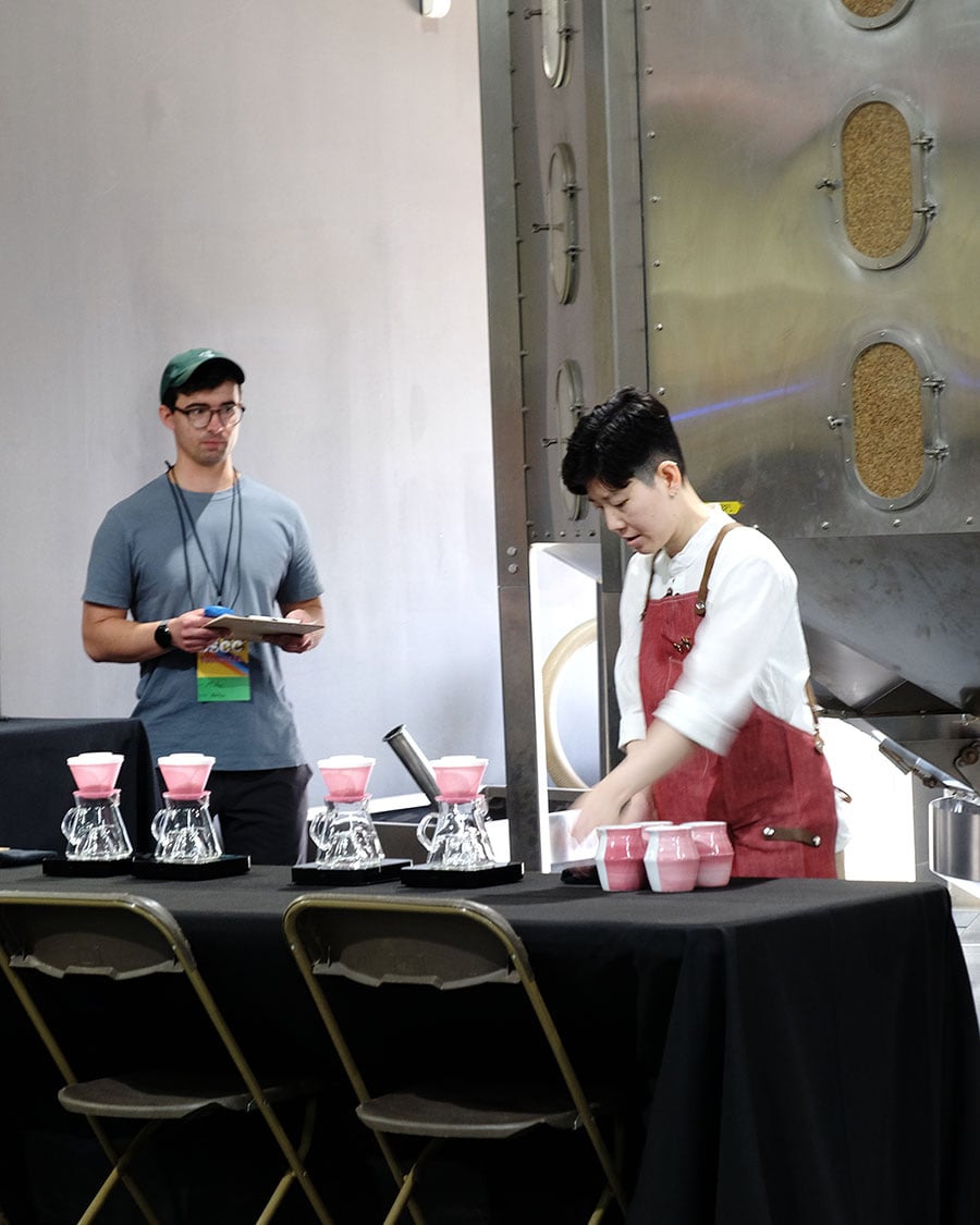 Peace setting up her USBrC table. A lineup of pink brewers and cups sits on a black tablecloth.